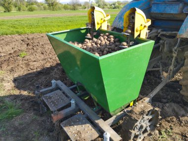 Bir tarlada mavi bir traktöre bağlı patates ekicisinin arka görüntüsü. Patates ekicisinin patatesle dolu büyük yeşil bir zıpzıpı var. Toprağa patates ekme mekanizması altında görünür..