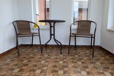 Yerde kahverengi fayanslar olan küçük bir terasta yuvarlak koyu bir masa ve metal çerçeveli iki hasır sandalye var.. 