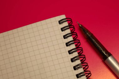 Pembe bir arka planda siyah spiral ciltli açık bir defter var. Yakınlarda siyah başlıklı gümüş bir tükenmez kalem bulunuyor. Not defteri beyaz, ince gri çizgiler kare biçiminde..