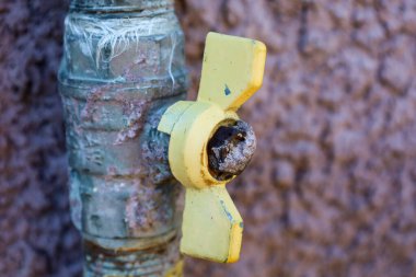 Düz olmayan bir duvarın arka planında sarı bir kapakçığı olan eski bir metal musluğa yakın çekim. Paslanma ve aşınma izleri görülebilir, bu da uzun vadeli kullanımı gösterir..