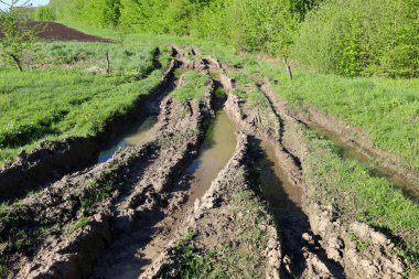 Çamurlu suyla dolu derin monotonlukları olan bozuk bir toprak yol yeşil bir tarladan geçiyor. Ağır makinelerin izleri ıslak zeminde açıkça görülebiliyor. Bu da yolculuk için dengesiz ve zor bir yol oluşturuyor..