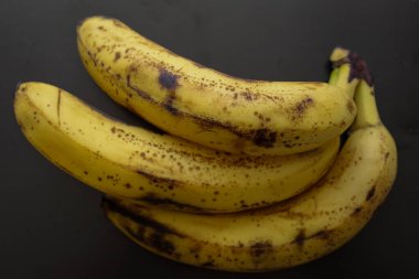 Üç olgun muz, koyu bir mat yüzeyinde üst üste yatar. Sarı kabuk çok sayıda kahverengi noktayla kaplıdır, bu da onların tüketime ya da pişirmeye hazır olduklarını gösterir..