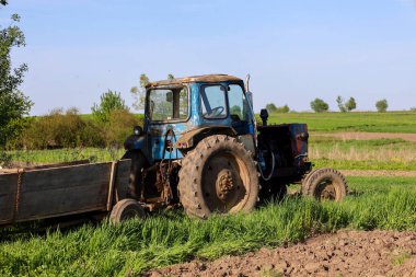 Eski mavi bir traktör yeşil bir tarlanın kenarında duruyor, kısmen kahverengi topraklarda, parlak bir gökyüzünün altında..