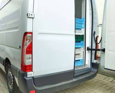 Açık kapıları olan beyaz bir minibüsün arkasında kısmen mavi, beyaz ve yeşil plastik kasalar var. Minibüs kaldırım taşlarının üzerine park edilmiş..