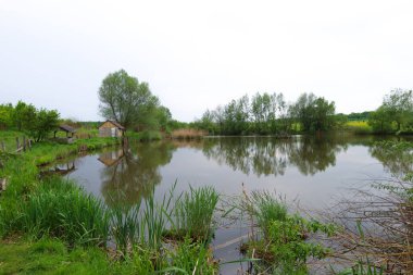 Gölün durgun suları ağaçlı yeşil kıyıları yansıtır. Kıyıda, gri çatılı ve tahta gölgelikli küçük ahşap bir yapı görülebilir..