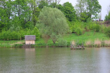 Gölün sakin su yüzeyi kıyıları çevreleyen yeşil ağaçları yansıtır. Bir kıyıda, gri çatılı küçük ahşap bir yapı görünür, ve diğer tarafta, ahşap bir platform kısmen suya batar..
