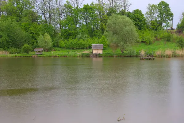Gölün durgun suları bulutlu gökyüzünü ve ağaçlı yeşil kıyıları yansıtır. Kıyıda, koyu renkli çatısı ve ahşap gölgeliği olan küçük açık renkli bir bina görülebilir..