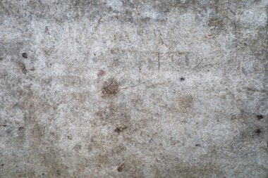 Gri beton yüzeyde aşınma ve çizik izleri var. Betonun dokusu pürüzlü, küçük taşlar ve girintiler var..