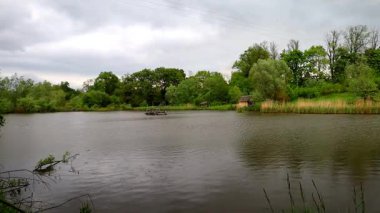 Yağmurlu havada küçük bir gölet. Damlalar sakin su yüzeyine düşer ve dalgalanmalar yaratır. Suyun kenarındaki yemyeşil bitki örtüsü nemle doludur. Atmosfer huzurlu ve taze..