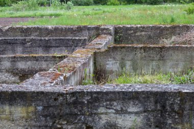 Beton bir temelin kalıntıları bulutlu bir gökyüzünün altındaki kırsal araziye bakan genç yeşil çimlerin arasından bakıyor. Geçmişin izleri doğanın yeniden doğuşuyla tezat oluşturuyor..