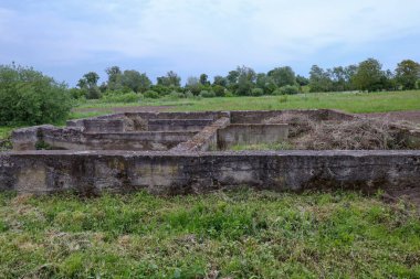 Eski bir yapının beton kalıntıları yeşil bir manzaranın ortasında çimlerle kaplıdır. Dokulu yüzeylerinde zaman ve çürüme izleri var..