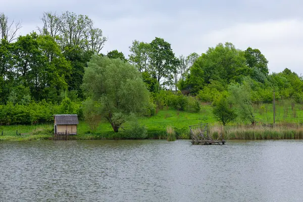 Yeşillik ve ağaçlarla çevrili sakin bir göl manzarası. Kıyıda küçük ahşap bir yapı görülebilir, suda bir iskelenin kalıntıları var. Bulutlu gökyüzü sahneye melankolik bir hava katıyor..
