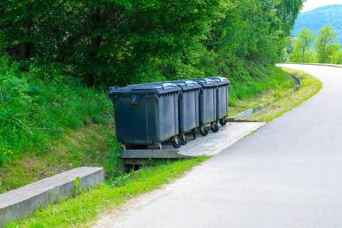 Yeşil bir orman zeminine karşı yol boyunca yerleştirilen bir dizi koyu çöp konteyneri, sorumlu atık yönetiminin önemini vurguluyor.