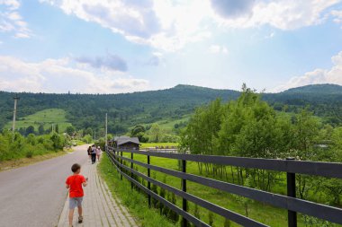 Urich, Ukrayna, - 08 Haziran 2024: Bir çocuk, yeşil bir tarlayı yoldan ayıran, siyah ahşap bir çitin yanında asfalt bir yol boyunca yürüyor.