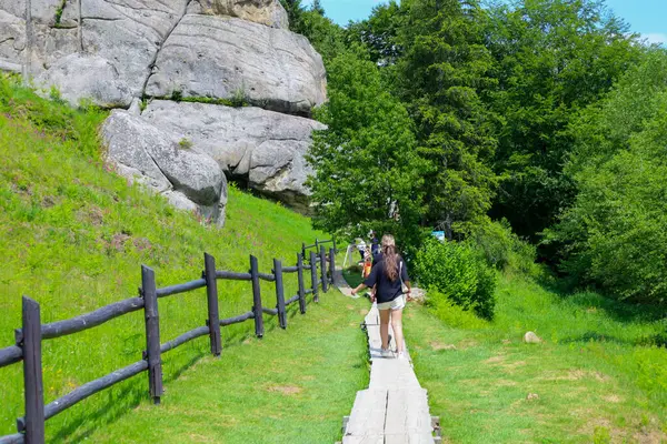 Urich, Ukrayna, - 08 Haziran 2024: Ahşap tahtalardan oluşan bir patika, kayalık kayalıklar ve gür ağaçlar tarafından çerçevelenmiş yeşil bir araziye çıkar. Gezginler doğada huzurlu bir yürüyüşün tadını çıkarırlar. 