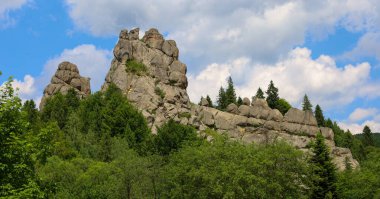 Yeşilliklerle kaplı güçlü kaya oluşumları sık bir ormanın üzerine yükselir ve etkileyici bir doğal manzara yaratır. Beyaz bulutlu açık mavi bir gökyüzü bu görkemli manzarayı tamamlıyor, vahşi doğanın güzelliğini vurguluyor..