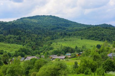 Geniş panoramik bir manzara, evleri olan rahat bir kırsal yerleşim bölgesinin üzerinde yükselen yeşil orman dağlarını gösteriyor. Hafif bulutlu açık bir gökyüzü sükunet ve doğal güzelliğin atmosferini tamamlıyor..