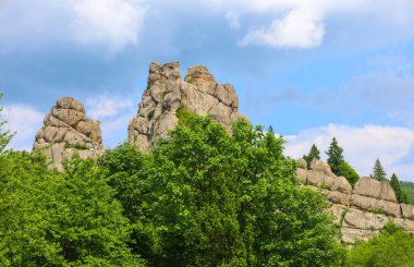 Yeşilliklerle kaplı güçlü kaya oluşumları sık bir ormanın üzerine yükselir ve etkileyici bir doğal manzara yaratır. Beyaz bulutlu açık mavi bir gökyüzü bu görkemli manzarayı tamamlıyor, vahşi doğanın güzelliğini vurguluyor..