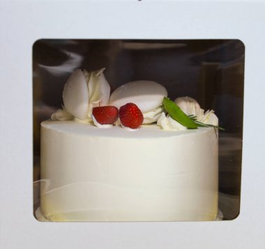 Beyaz kremalı, çilek, krema ve badem kurabiyeleriyle süslenmiş zarif bir pasta, şeffaf pencereli bir kutuya dikkatlice paketlenir. Bu tatlı ideal bir hediye ya da ikram olarak nakledilmeye hazır..