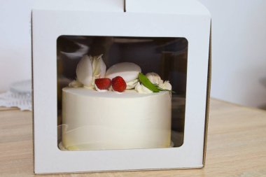 Beyaz kremalı, çilek ve kremayla süslenmiş düzgünce paketlenmiş bir pasta, bir karton kutunun penceresinden sergilenir..