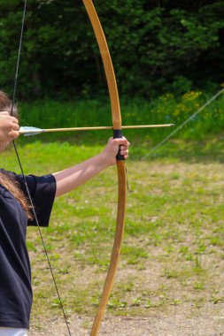 Bir okçunun elleri, yeşil çimen ve ağaçlardan oluşan bir arka planla ateş etmeye hazırlanan ahşap bir yayın ipini geri çeker. Bu an, antik bir sporda odaklanmayı ve beceriyi gösteriyor..