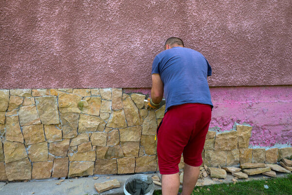 Мужчина кладет облицовочный камень на стену дома, тщательно работая над каждым элементом. Процесс создания прочного и эстетичного цоколя отражает мастерство и внимание к деталям в строительных работах.