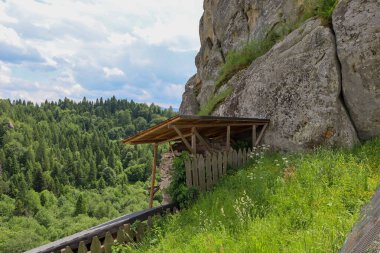 Yüksek doğal oluşumlara sahip kayalık bir arazi, yanında bir tepe örtüsü ve çitin bulunduğu basit bir ahşap yapı. Çekim, vahşi doğa ve insan müdahalesinin tarihsel ya da turistik bölgeleri göstermektedir..