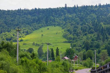 Ormanlarla kaplı yeşil Carpathian dağlarının geniş bir manzarası var, açıklıklar ve aşağılara yayılmış bir köy, dolambaçlı bir yol ve elektrik direkleriyle.