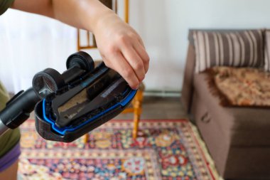 Bir kadın eli elektrikli süpürgeyi baş aşağı tutuyor, kullandıktan sonra iplik ve tozla gösteriyor. Arka planda, kanepesi ve halısı olan bir oda görülüyor, temizlik sürecini vurguluyor..