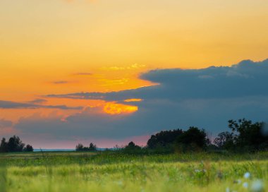 Yeşil bir tarlanın üzerinde büyüleyici bir günbatımı görünür. Işıklar kara bulutları delip geçer ve altın kırıklar yaratır. Ufkun altında, gökyüzü narin pembe renkler alır..