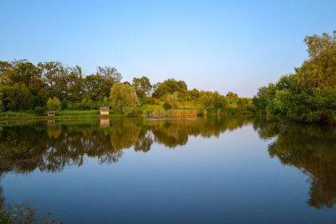 Sakin bir göl bulutsuz bir gökyüzünü ve yeşil ağaçları yansıtır ve harmonik bir yaz manzarası yaratır. Sahilde küçük bir ev görülebiliyor, bu huzurlu resme rahatlık katıyor..