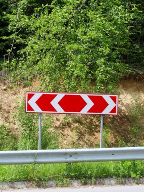 Yol kenarında duran parlak kırmızı-beyaz bir simge keskin bir virajı işaret ediyor. Arka planda, yeşil orman bitki örtüsü ve toprak yamacı görülebiliyor ve doğal çevre ile karayolu altyapısı arasında bir tezat oluşturuyor..