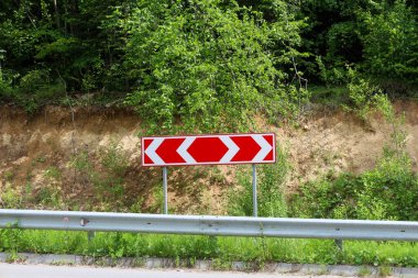 Yol kenarında keskin bir dönüş olduğunu gösteren parlak bir yol işareti var. Arkasında yeşil ağaçlar ve toprak seti doğal bir zemin oluşturur..