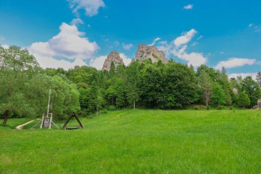 Bu manzara çekimi, bir ormanın üzerinde yükselen görkemli kayaları ön planda mavi bir gökyüzünün altında canlı yeşil bir tarlaya sahiptir..