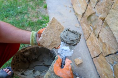 Eldivenli bir işçinin elleri, yerleştirmeden önce dekoratif taşlara dikkatlice havan topu sürüyor. Bu süreç, titiz duvar bitirme, doku ve doğal bir görünüm eklemeyi gösteriyor. İnşaat ve yenileme projeleri için mükemmel..