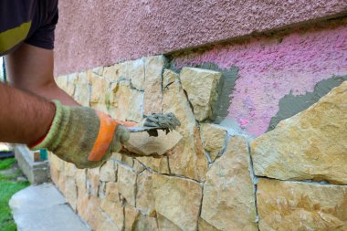 Eldivenli bir işçi bir binanın ön cephesine havan topu kullanarak dekoratif taşlar koyuyor. Bu resim, tasarım çalışmalarının bitirilmesi, inşaatın tamamlanması ve doğal malzemelerin kullanımının sürecini gösteriyor..