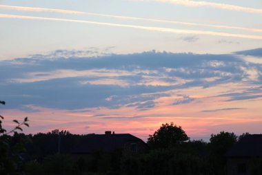 Renkli bir günbatımına karşı ev ve ağaç siluetleriyle huzurlu bir akşam manzarası. Gökyüzü pembe, turuncu ve mavi tonlarıyla parıldıyor. Huzur ve sükunet ortamı yaratıyor..