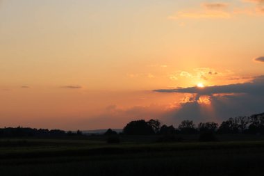Kırsal bir arazide büyüleyici bir günbatımı, sıcak ve dingin bir ışık saçıyor. Güneş ışınları bulutları delip geçer, ufku aydınlatır ve sahneye dram katar.. 