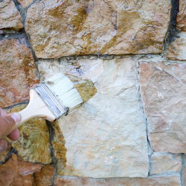 Bir zanaatkârın eli, bir fırçayla taş bir duvara koruyucu bir solüsyon uygular ve rengini değiştirir. Bu süreç ev bakımını ve cephe yenilemesine yönelik profesyonel yaklaşımı yansıtıyor.