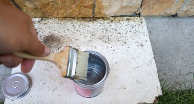 Bir el, metal bir solüsyon tenekesine fırça batırır ve taş bir duvarın yanında çalışmaya hazırlanır. Çevredeki yüzey kirlidir. Tamir ya da inşaat sürecini gösterir. İşçiliğin gerçeklerini vurgular..