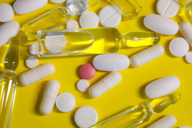 Parlak sarı arka planda, çeşitli tıbbi hazırlıklar düzenlenmiştir: beyaz yuvarlak ve dikdörtgen hapların yanı sıra sarı sıvı cam ampuller.