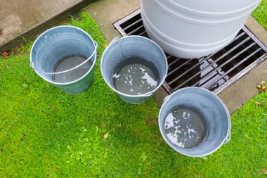 Yağmur suyu dolu üç galvanizli kova, kanalizasyon ızgarasının yanındaki yeşil çimlerde değerli doğal kaynakları topluyor. Bu sahne, ev ihtiyaçlarını karşılamak için yağmur suyunu kullanmanın ekolojik bir yaklaşımını gösteriyor. 