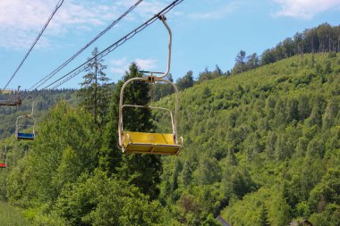 Boş sandalyeler, mavi gökyüzünün altındaki bir dağ yamacındaki yoğun yeşil bir ormanın üzerinde hareket ediyor. Bu pitoresk yaz manzarası dağlarda etkin bir eğlence ve doğanın keyfine davet ediyor..