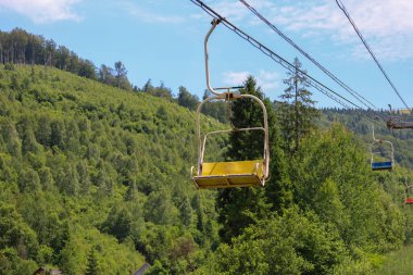 Yoğun bir ormanla kaplı, teleferik hatlarının geçtiği bir dağ yamacının manzarası. Açık yaz gökyüzünün altında, bu manzara gezmeyi ve dağların doğal güzelliğinin tadını çıkarmayı davet eder..
