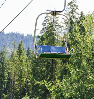 Metal bir çerçeve ve mavi-yeşil bir koltuktan oluşan bir teleferik havada asılı, yoğun bir kozalaklı ormanın üzerinde uzak dağlara ve mavi bir gökyüzüne doğru hareket ediyor..