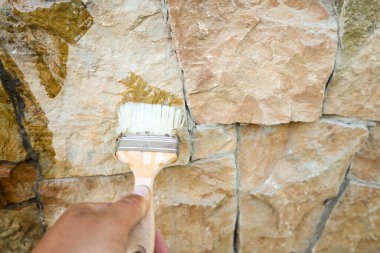 Bir el, taşın doğal dokusunu ve rengini vurgulayarak, bir taş duvara fırçayla sıvıyı dikkatlice uygular. Ön cephe bakım ve tedavi süreci ilginç bir görsel aksan yaratır..