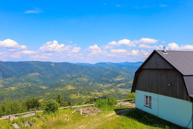 Yeşil dağların panoramik manzarasını sunan bir yamaçta, gable çatısı ve açık renkli duvarları olan kırsal bir ev yer alıyor. Beyaz bulutlu açık mavi bir gökyüzü sakin ve resimsel manzarayı tamamlıyor..