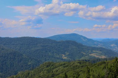 Fotoğraf, sonsuz ormanların sayısız tepe ve vadiyi kapladığı ve çok katmanlı yeşil bir rahatlama yaratan geniş bir dağ sırasını tasvir ediyor. Pofuduk bulutlu mavi bir gökyüzü manzaraya huzur verir..
