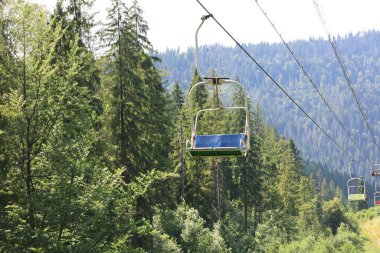 Bu fotoğraf bir dağ yamacında sık bir ormanın üzerinden geçen bir asansörü gösteriyor. Asansörün sandalyeleri boş, sükunet ve beklenti hissi yaratıyor..
