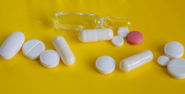 Açık bir tıbbi ampul, çeşitli haplar ve kapsüller içeren yatay bir bileşim, parlak sarı bir arka plana saçılmış..